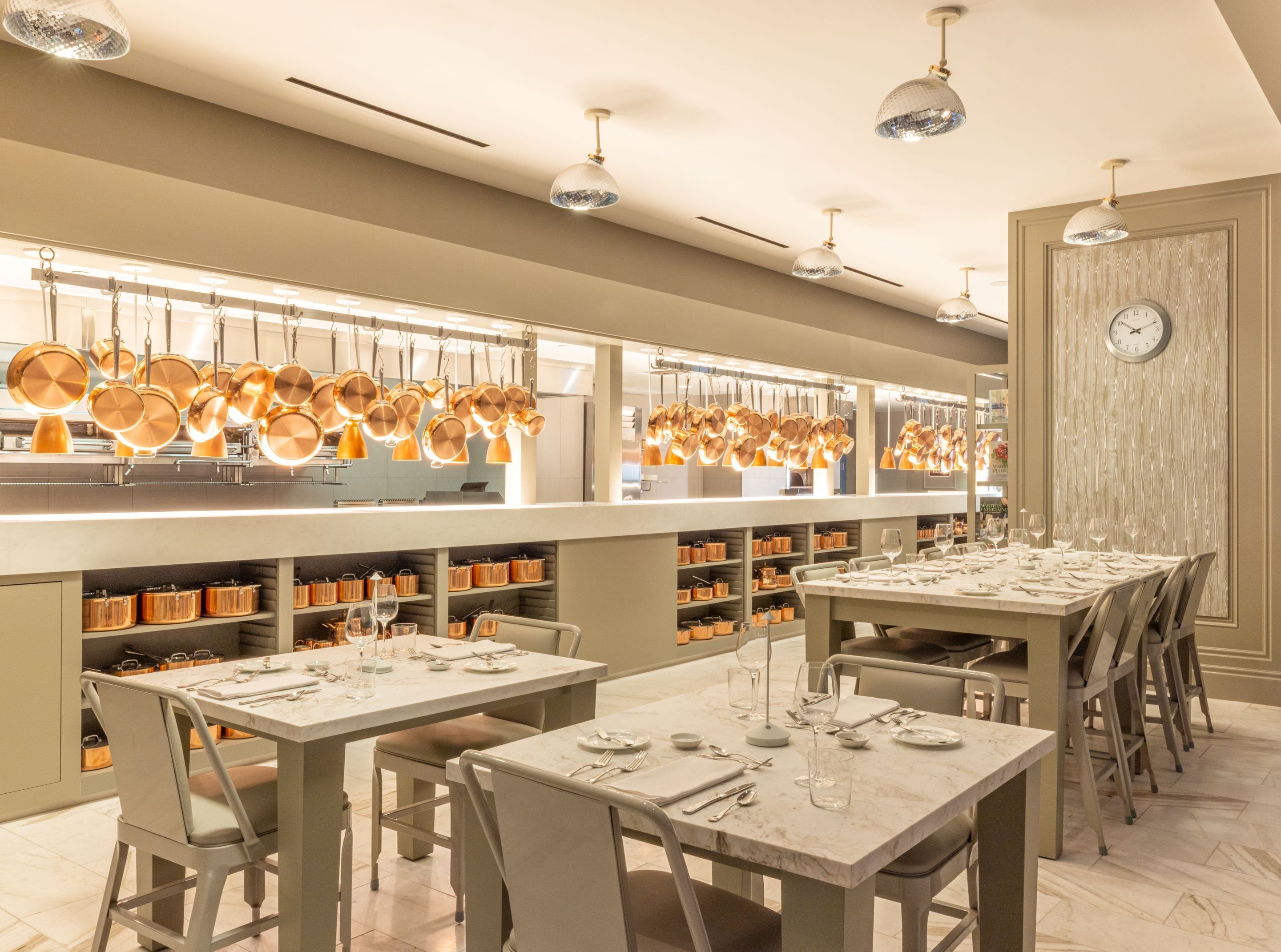A modern restaurant with a long counter displaying copper cookware, neatly arranged tables with cutlery, and a clock on the wall.