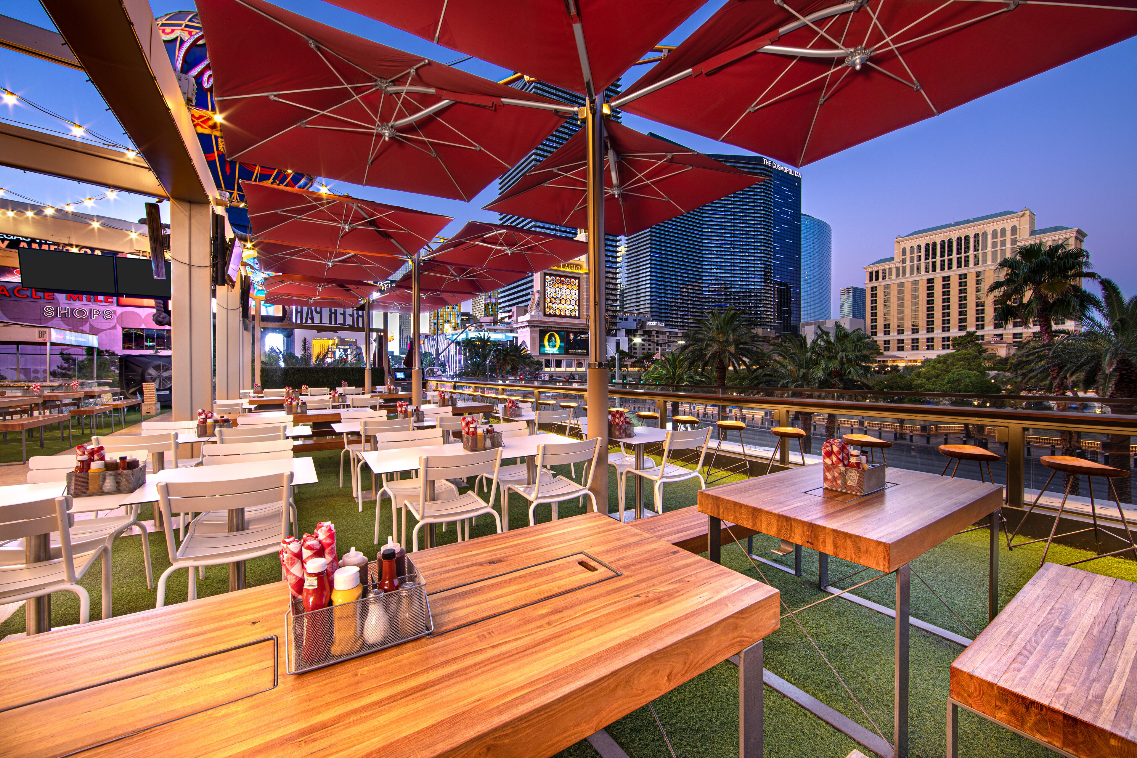 An outdoor dining area with tables and chairs, red umbrellas, and a view of the city skyline.