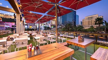 An outdoor dining area with tables and chairs, red umbrellas, and a view of the city skyline.