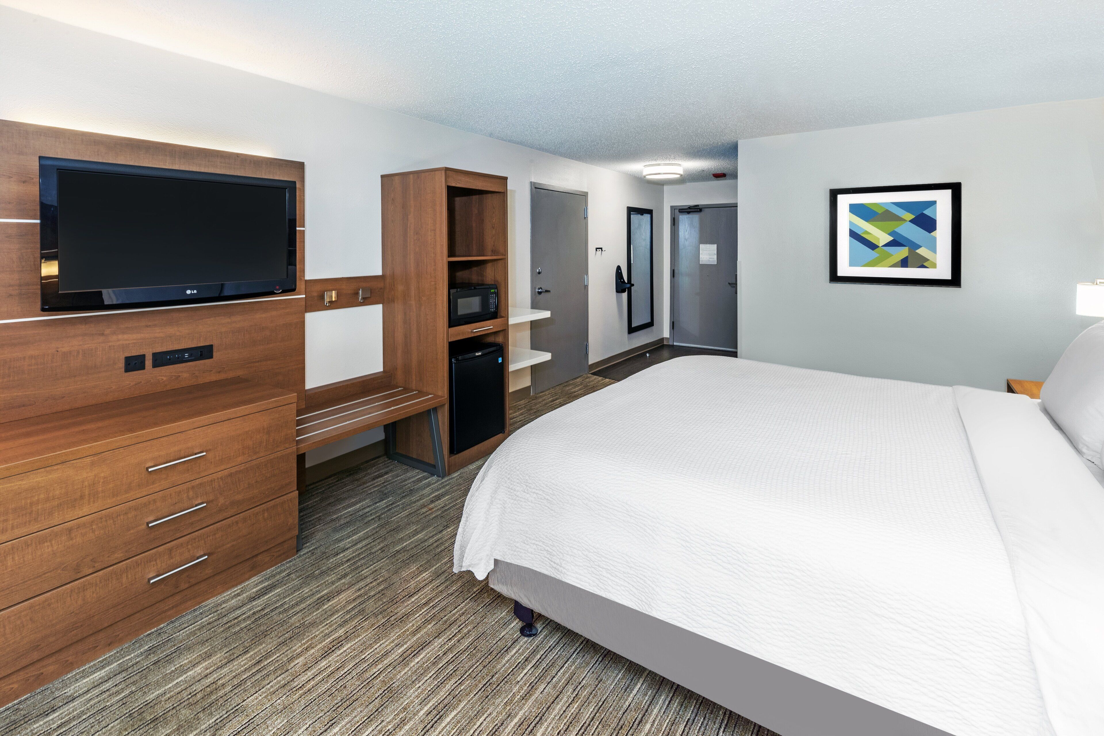 A hotel room with a bed, a TV, a desk with a microwave, a wardrobe, and a door.