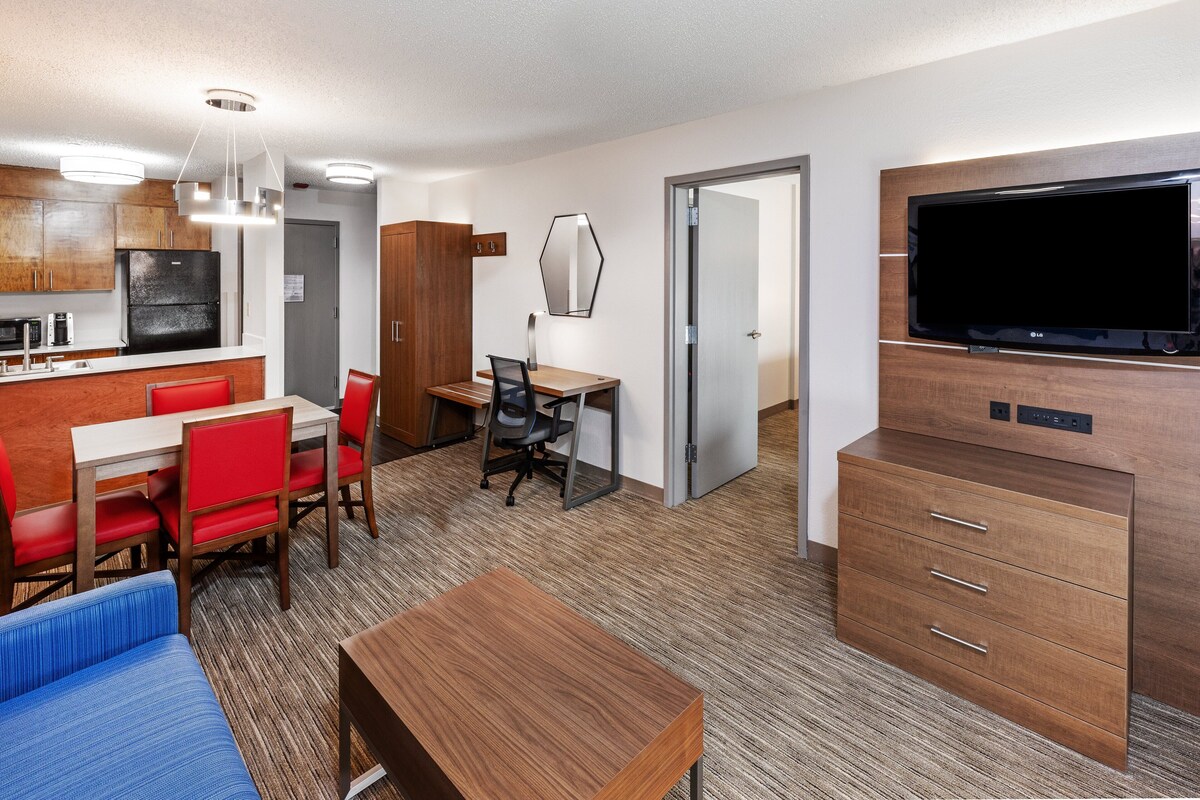 A compact hotel room with a kitchenette, a dining area with red chairs, a blue sofa, a wooden desk with a chair, and a TV mounted on the wall.