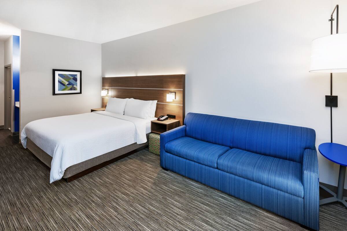 A hotel room with a bed, a blue sofa, a nightstand with a lamp, and a framed picture on the wall.