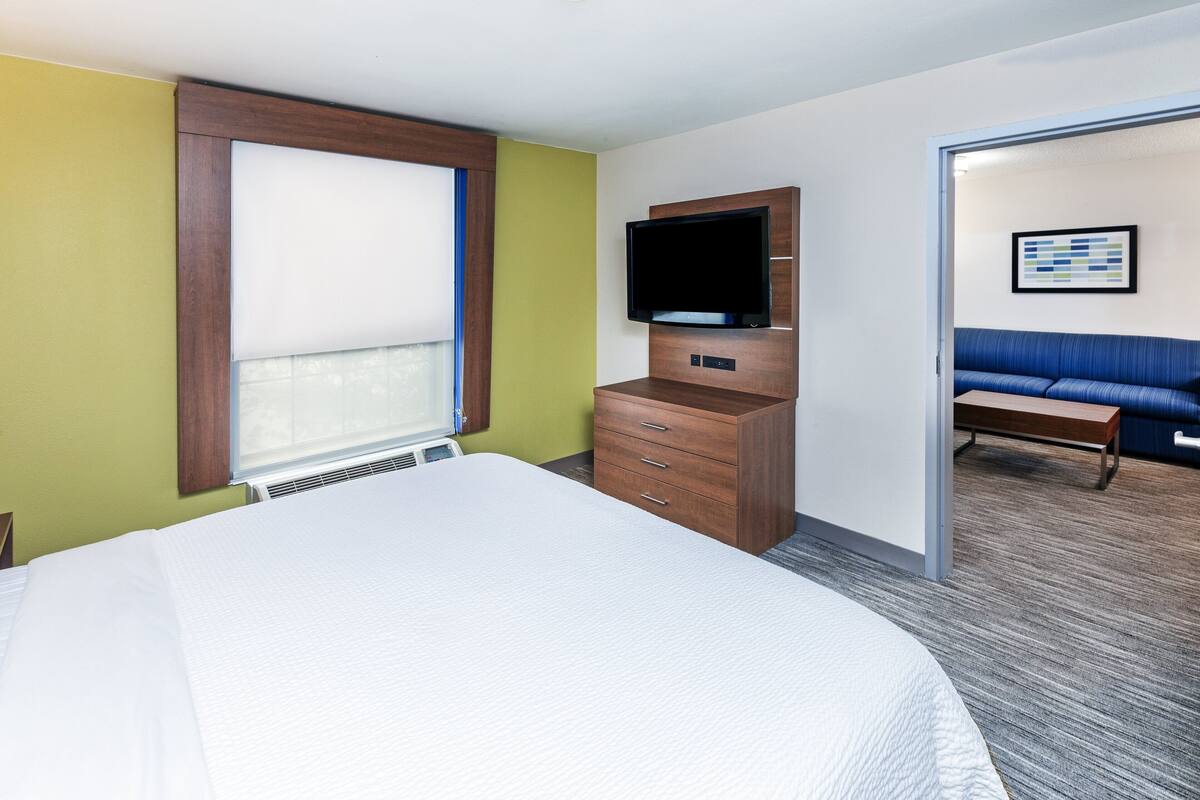 A hotel room with a large bed, a television on a wooden stand, a blue sofa, and a window with blinds.