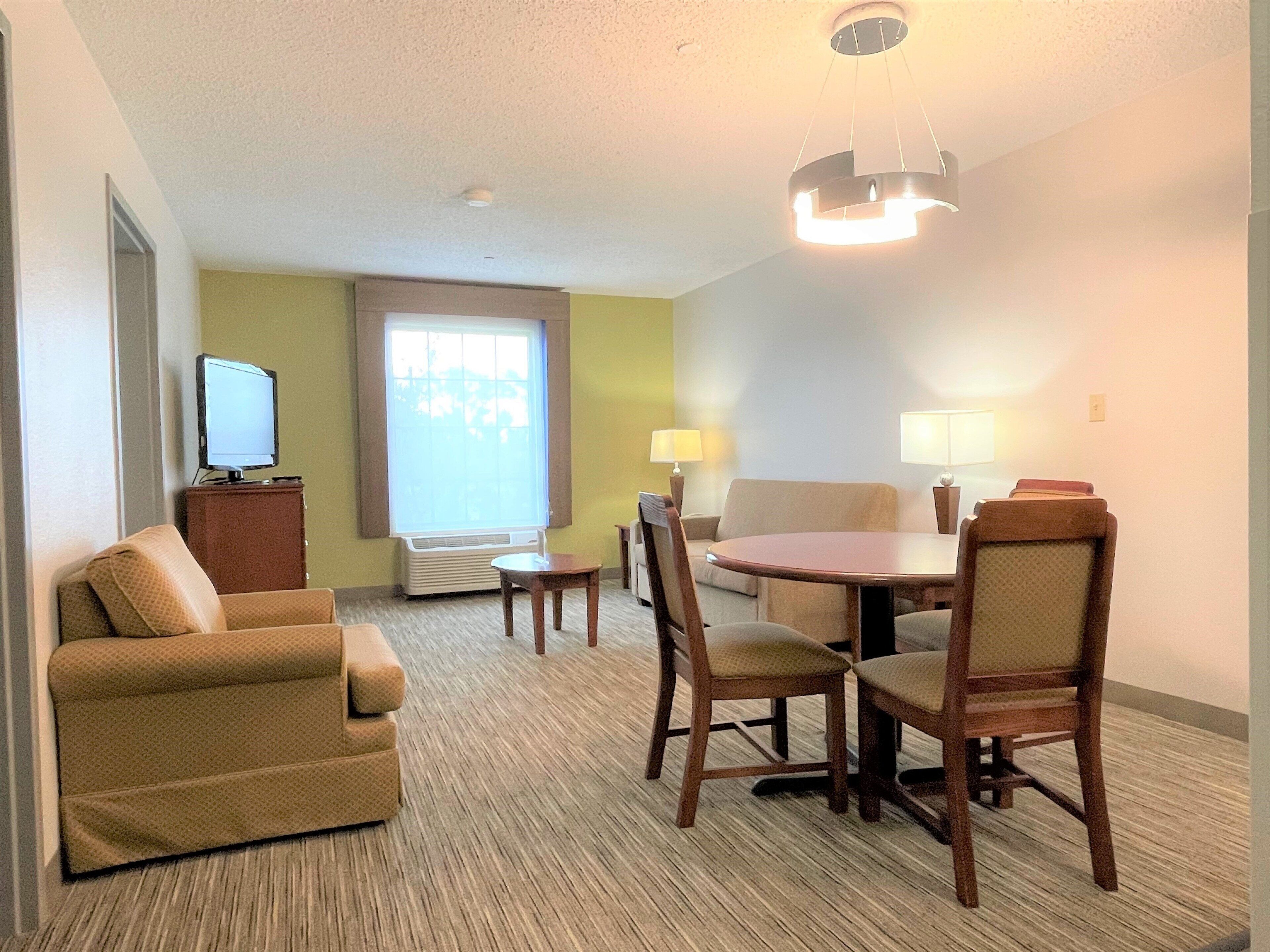 A hotel room with a sofa, a dining table with chairs, a television, and a window with blinds.