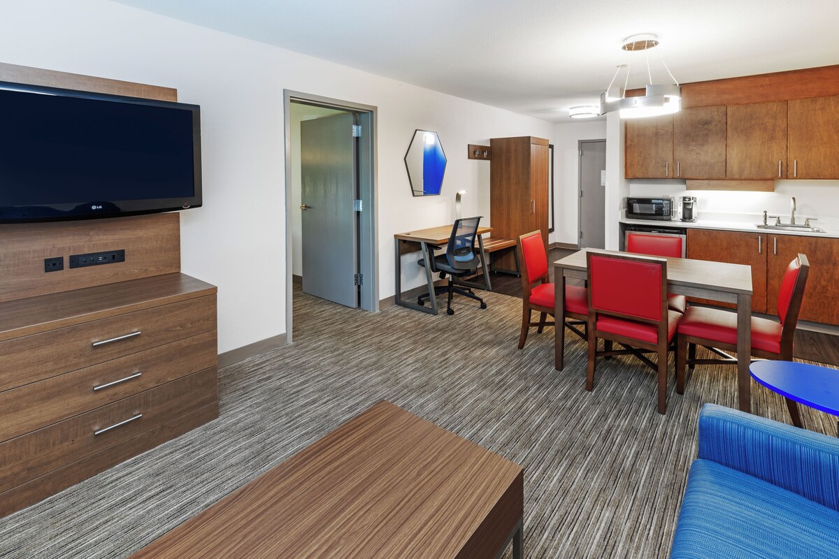 A modern hotel room with a flat-screen TV, wooden furniture, a dining area with red chairs, and a kitchenette.