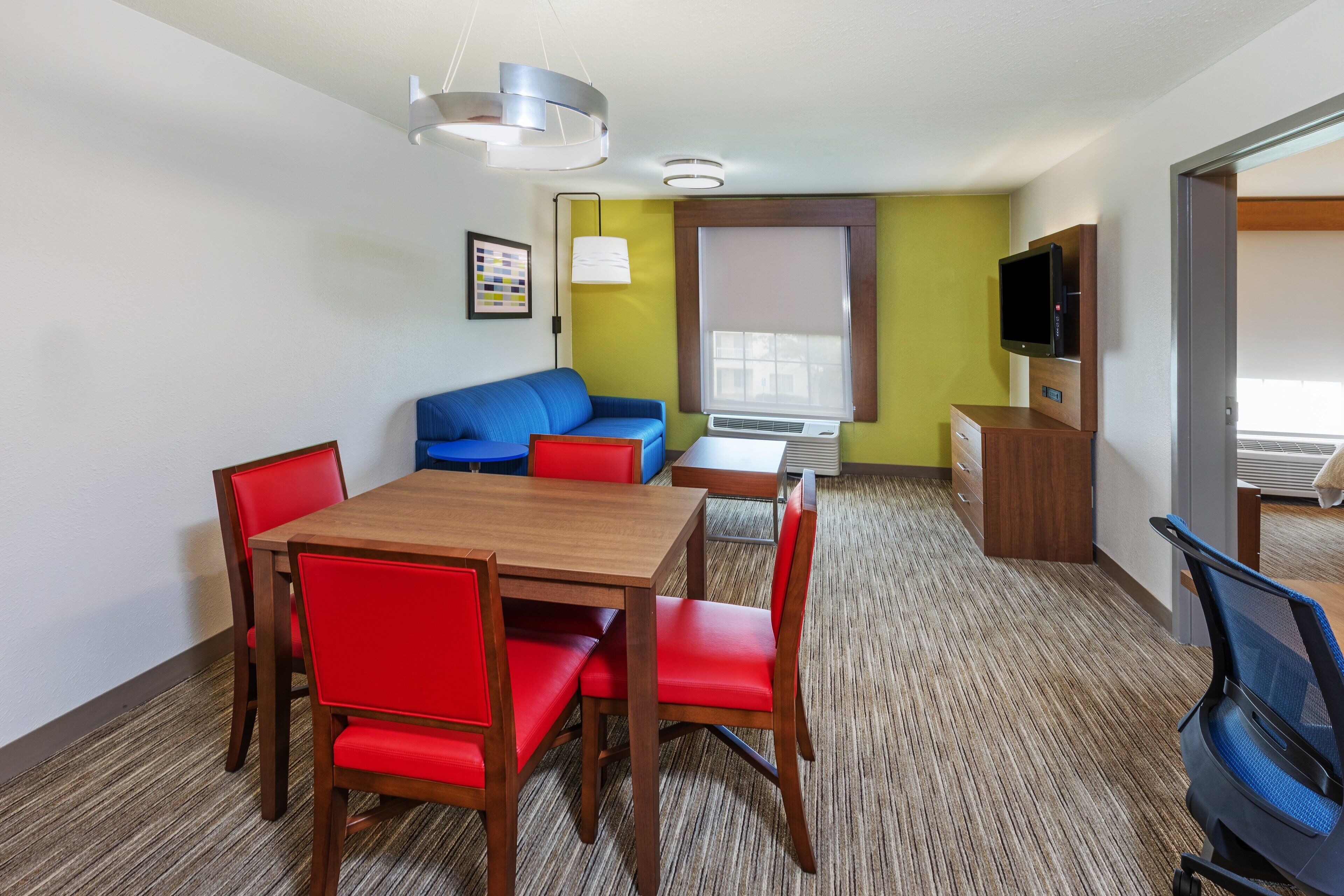 Habitación de hotel con mesa de comedor de madera, sillas rojas, sofá azul, televisor y ventana con persianas.
