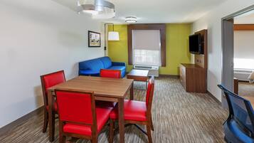 Habitación de hotel con mesa de comedor de madera, sillas rojas, sofá azul, televisor y ventana con persianas.