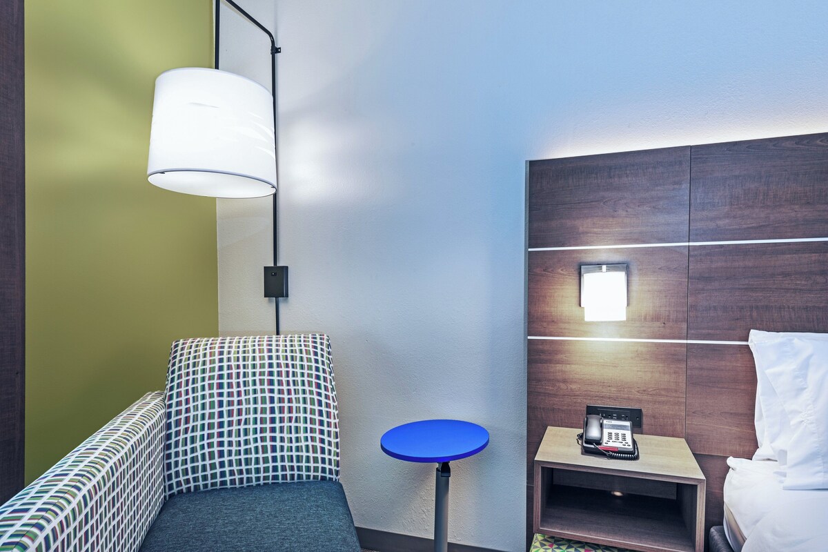 Habitación de hotel con una cama, una mesita de noche con un teléfono, una mesa redonda azul, un sofá estampado y una lámpara colgante.