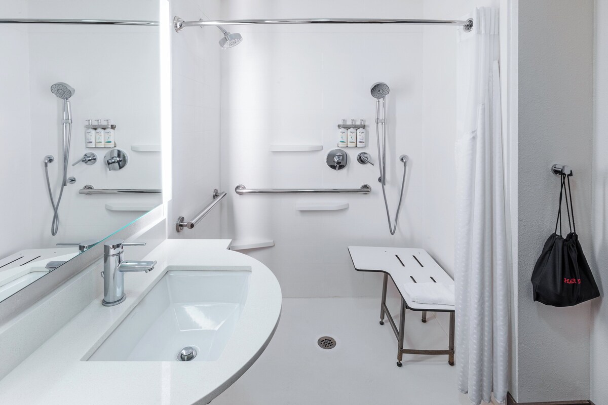 Un baño moderno con un lavamanos con recinto de vidrio, una ducha con cabezal de ducha manual y una barra de apoyo.