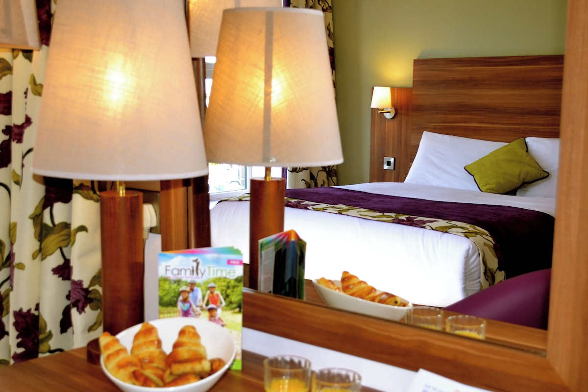 A hotel room with a wooden headboard, a bed with a purple bedspread, a green pillow, and a bedside table with a bowl of croissants, glasses of orange juice, and a magazine.