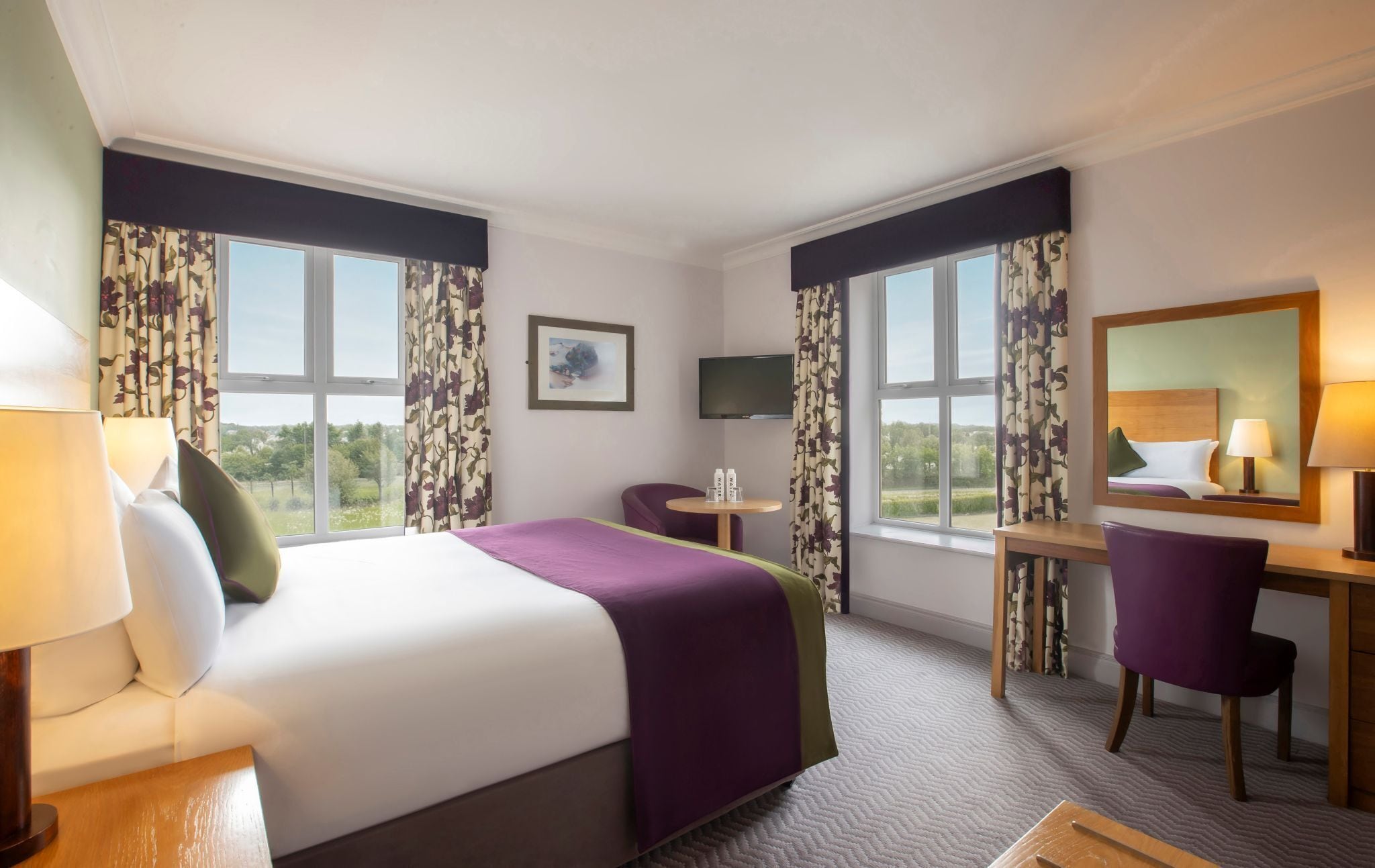 Ein Hotelzimmer mit einem Bett, einem Schreibtisch, einem Sessel, einem Fernseher und großen Fenstern mit Vorhängen.