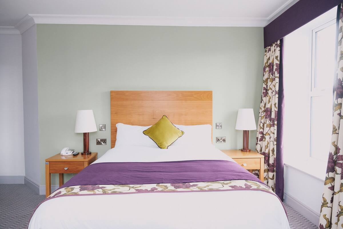 Hotellihuone, jossa on suuri sänky, kaksi yöpöytää valaisimineen, puhelin ja ikkuna, jossa on kukalliset verhot.