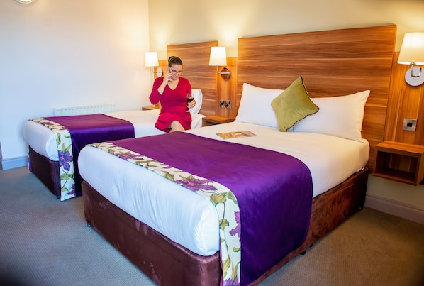 A hotel room with two beds, each with a wooden headboard and a purple bedspread. There are bedside lamps and a nightstand with a lamp on each side.