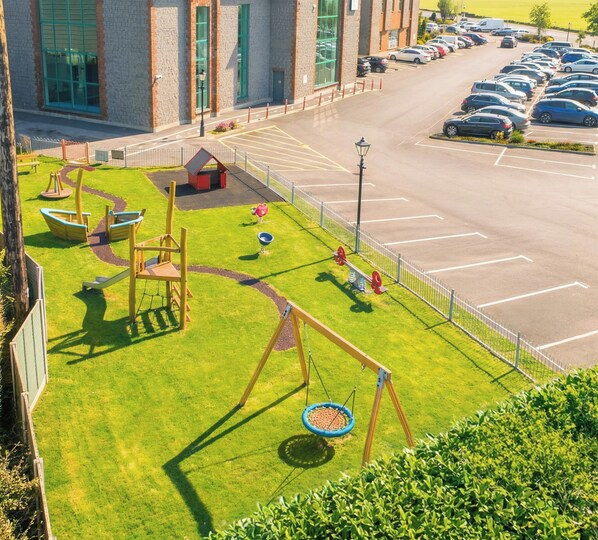 A playground with swings and a slide, a parking lot with cars, and a building with large windows. - Maldron Hotel, Oranmore Galway (Oranmore)