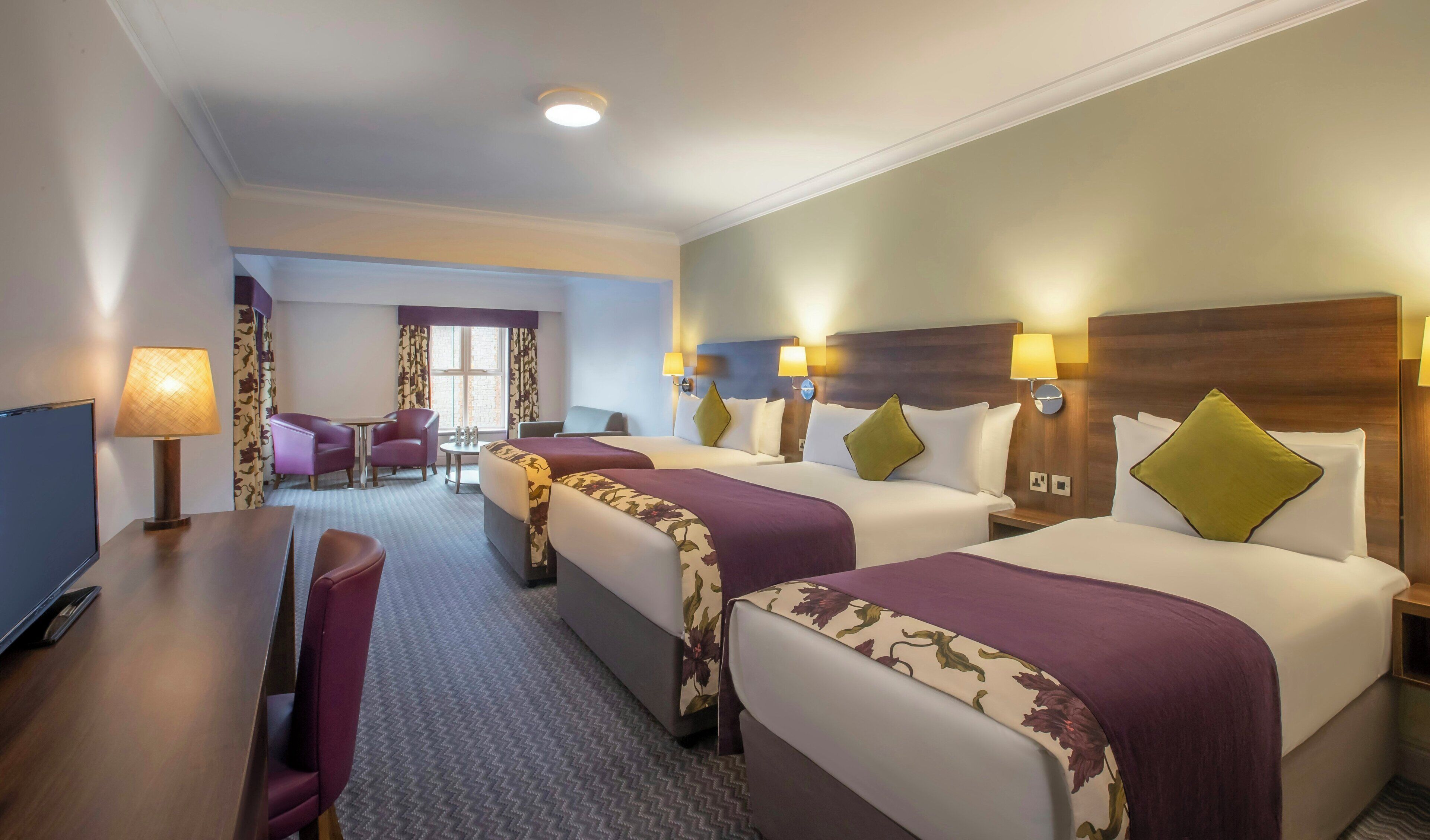 A hotel room with three beds, a desk, a chair, a TV, and a view of the hotel lobby.