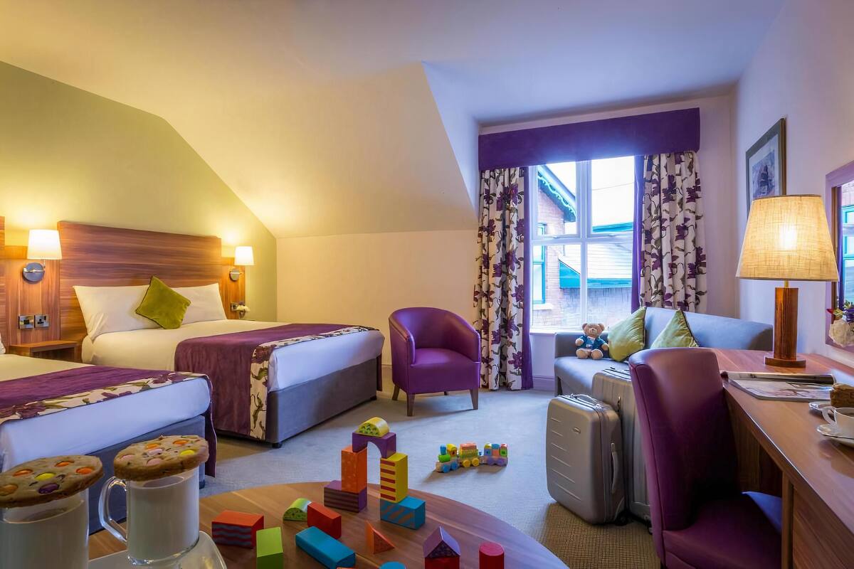 Hotellihuone, jossa on kaksi sänkyä, violetti nojatuoli, työpöytä lampun kanssa, pehmolelu ja ikkuna, jossa on verhot.