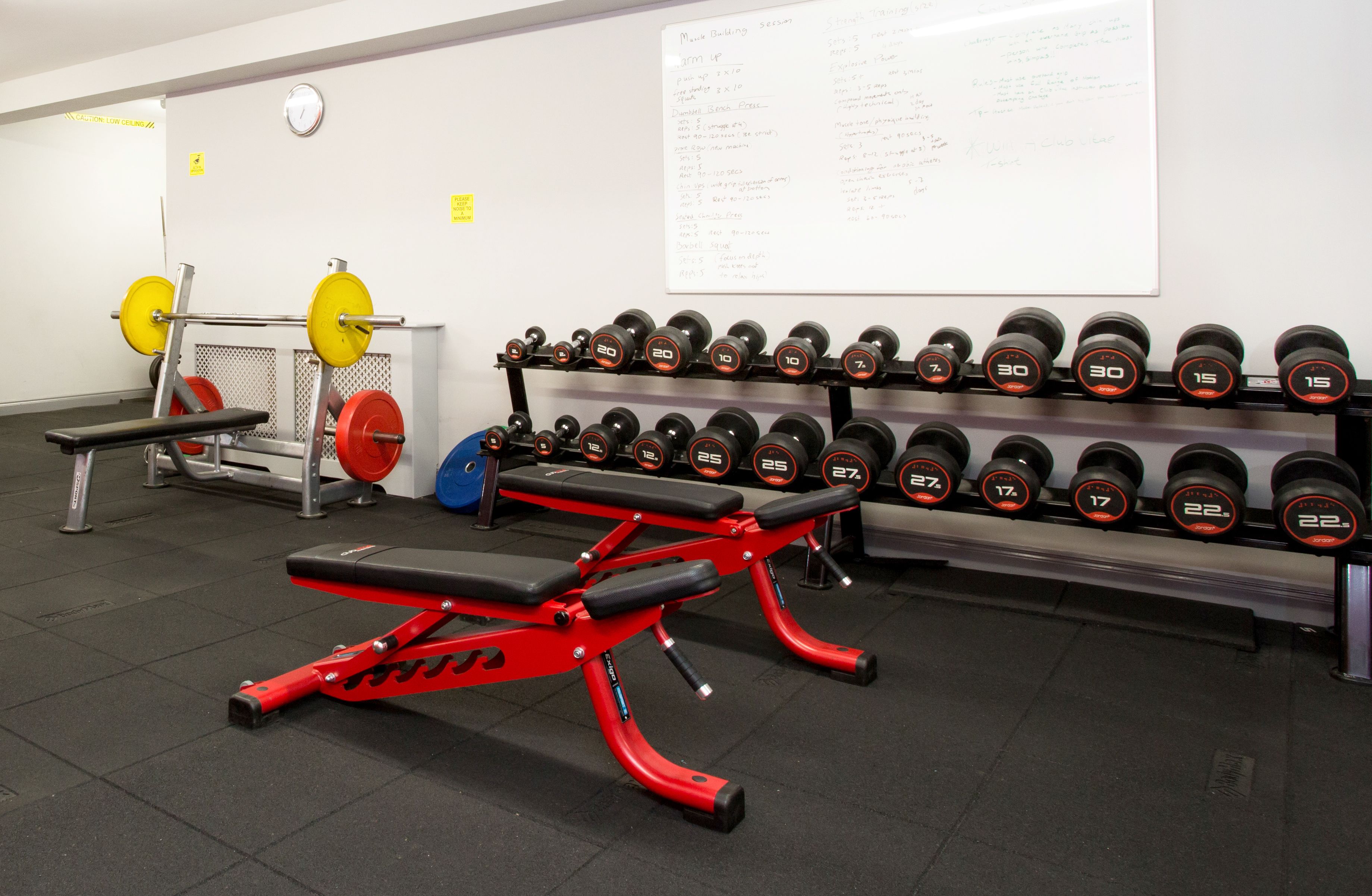 A gym with weightlifting equipment, a bench, and a wall with a list of exercises.