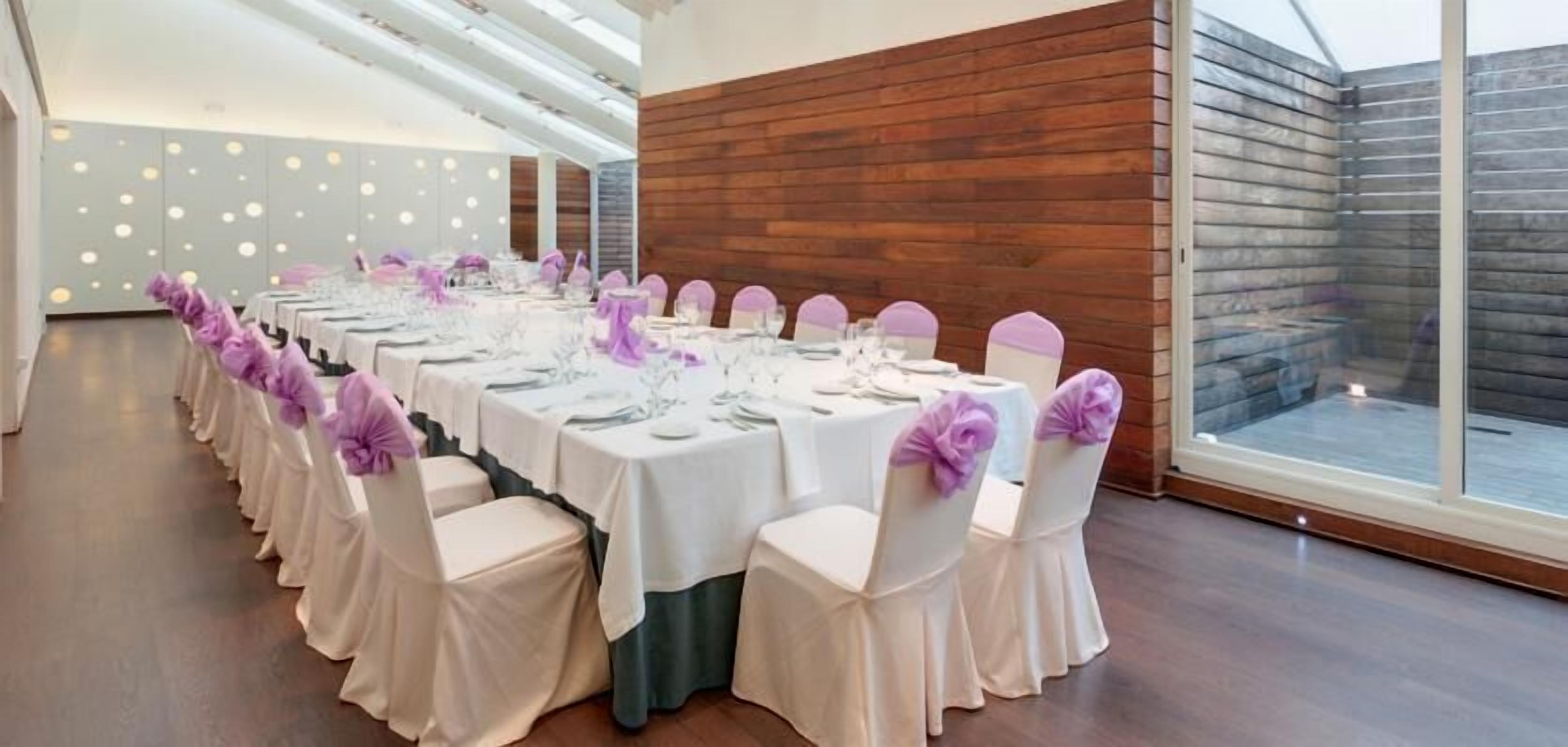 Una larga mesa dispuesta para un evento formal, con manteles blancos y cintas moradas, rodeada de sillas con cintas a juego.