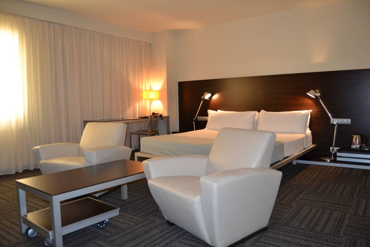 Una habitación de hotel con una cama, dos sillones blancos, una mesita, un escritorio y una lámpara.