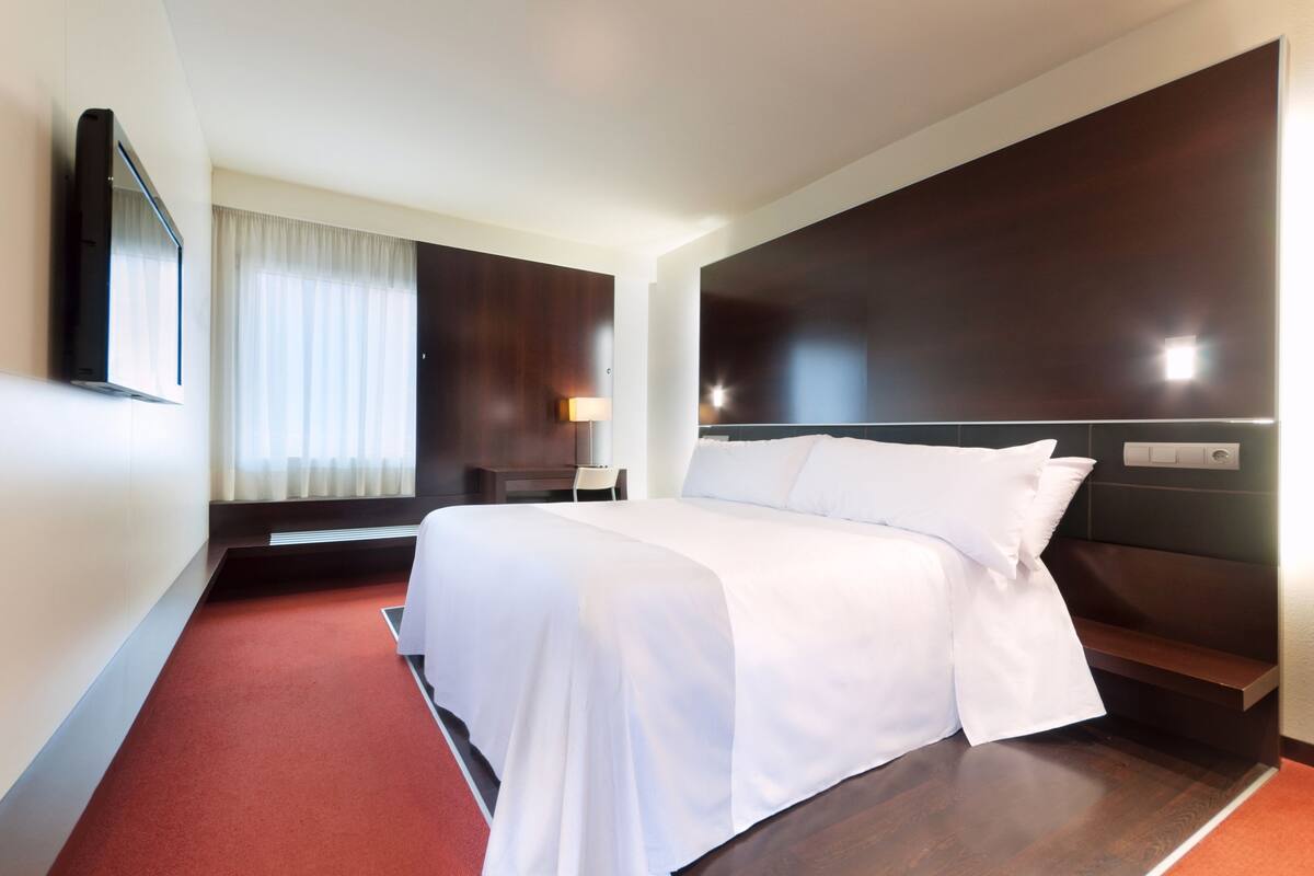 Una habitación de hotel con una cama grande, un televisor de pantalla plana en la pared, una mesita de noche con lámpara y una ventana con cortinas.