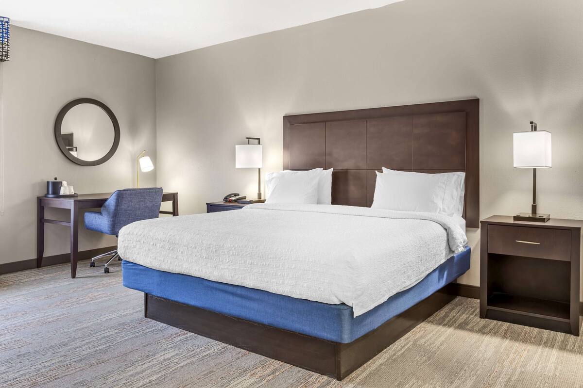 A hotel room with a large bed, a desk with a chair, a round mirror, and two bedside tables with lamps.