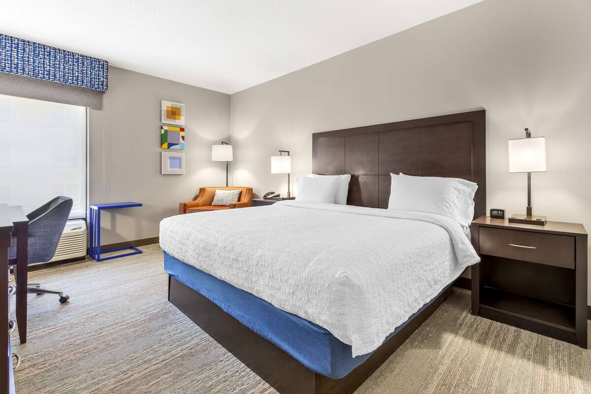 A hotel room with a large bed, a desk, a chair, and a nightstand with a lamp.