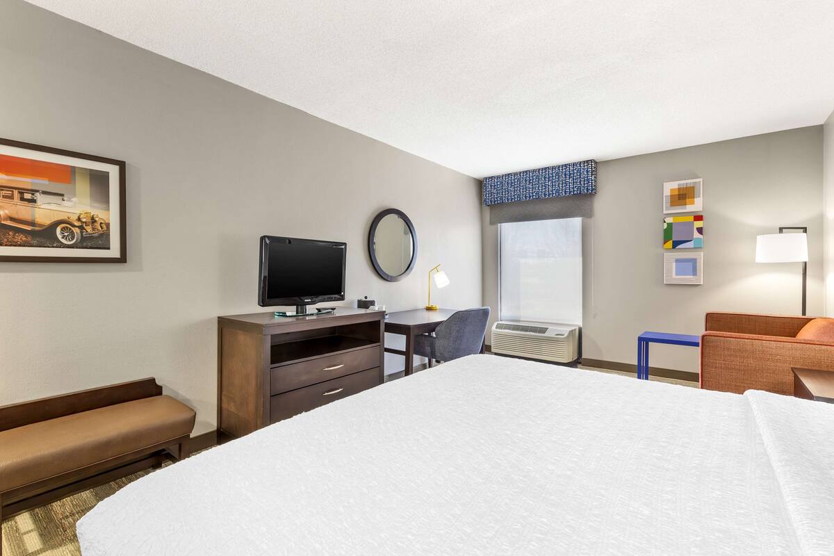 A hotel room with a large bed, a desk with a computer, a television, a window with blinds, a bench, and a wall-mounted mirror.