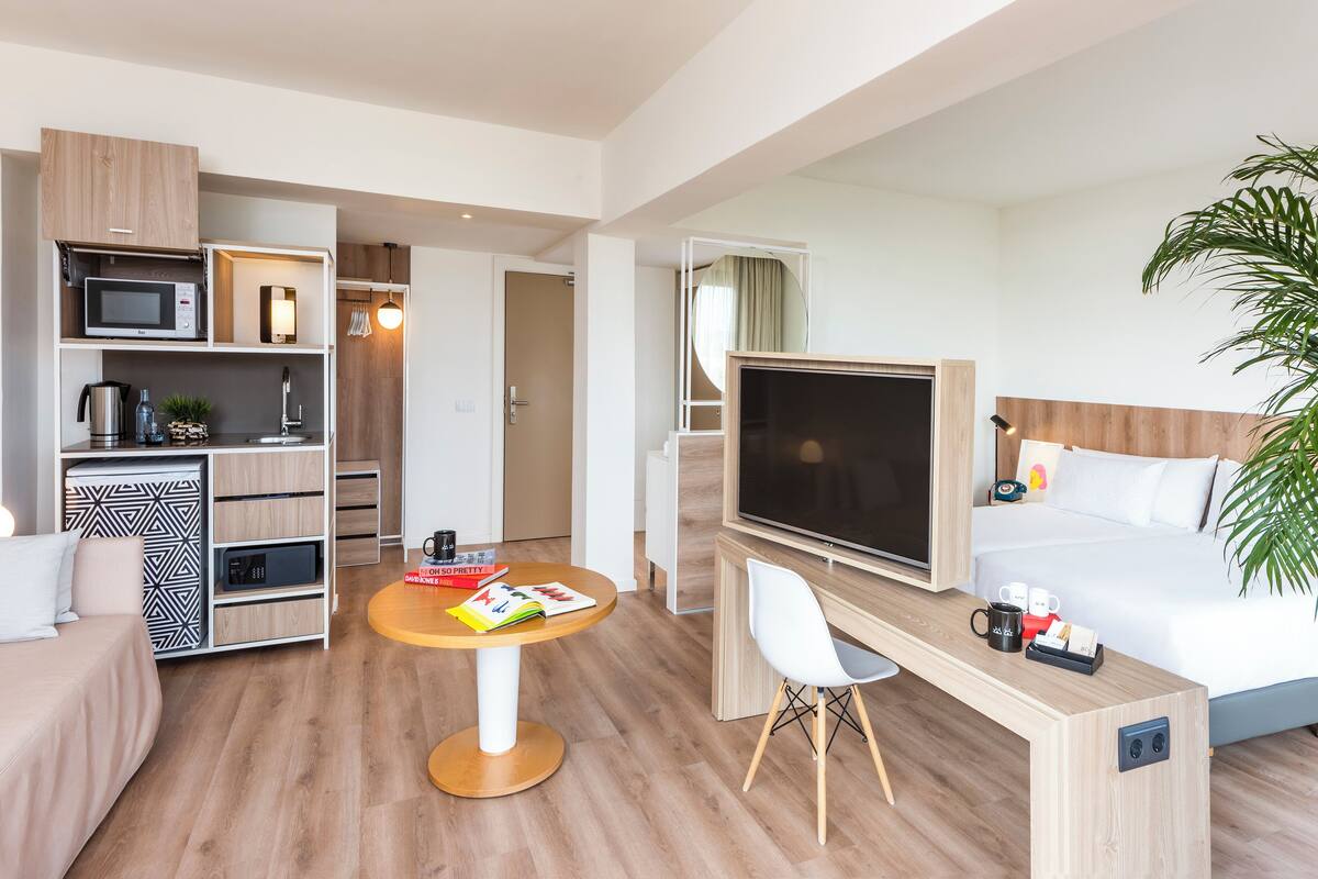 Una habitación de hotel moderna con una cama, un televisor, un escritorio con una silla, un microondas, una cafetera y una pequeña mesa con libros y una taza.