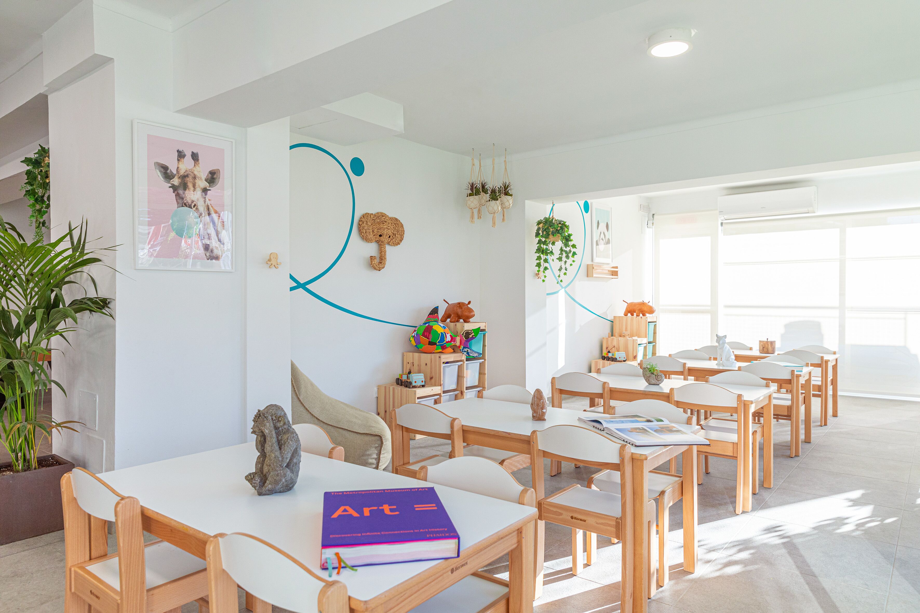 Ein helles, modernes Klassenzimmer mit weißen Wänden, Holztischen und Stühlen. An den Wänden befinden sich Lernplakate und ein Blumentopf.