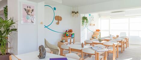 Uma sala de aula clara e moderna, com paredes brancas, mesas e cadeiras de madeira. Há cartazes educativos e um vaso de planta nas paredes.