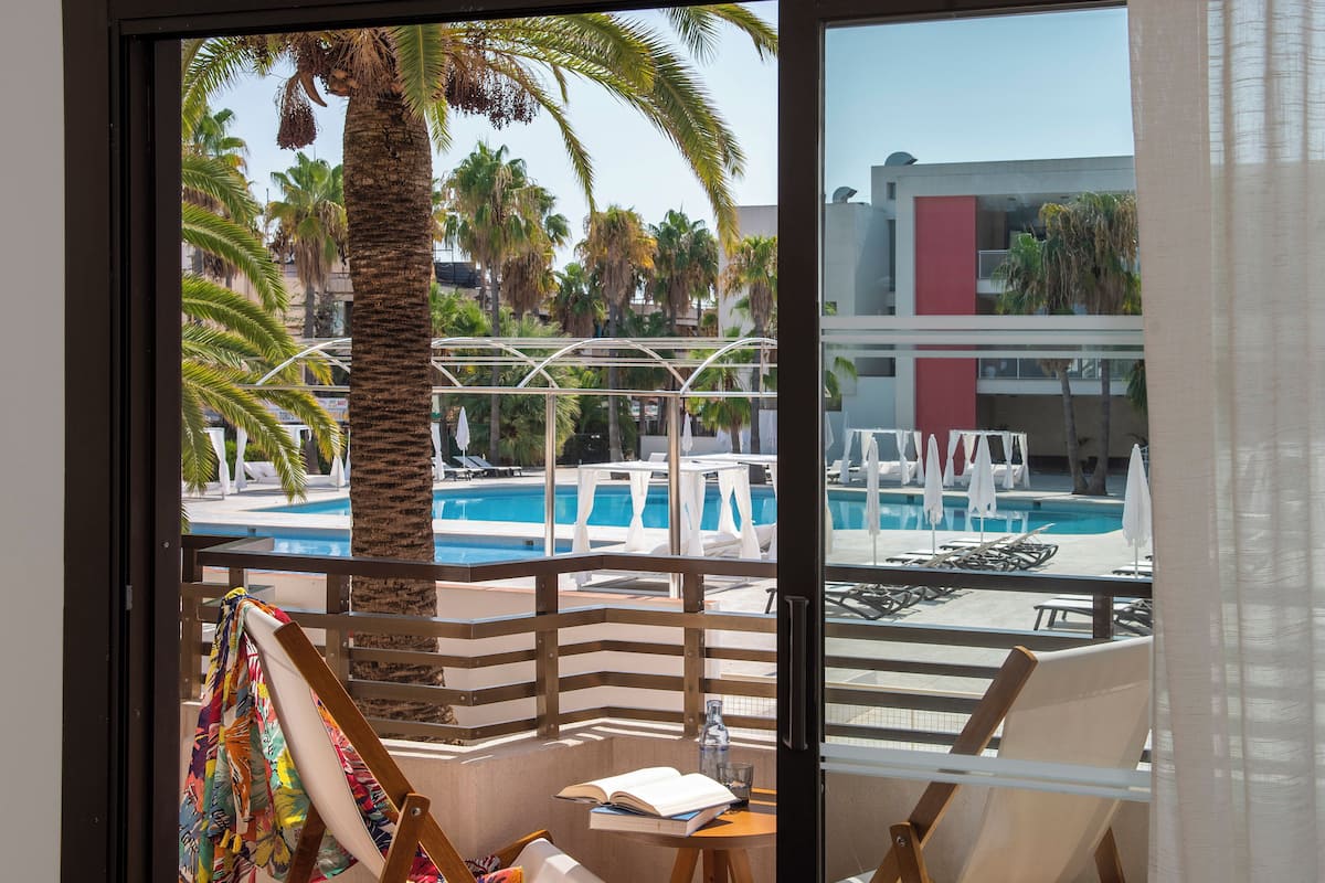 Un balcon avec vue sur la piscine, comprenant des chaises longues, une table avec un livre et un verre, et un palmier.