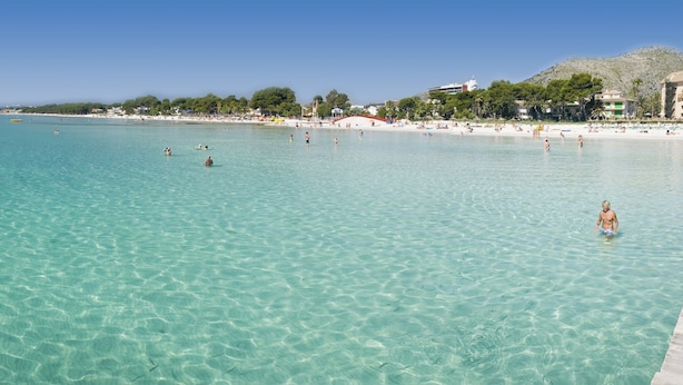 Une plage avec une eau turquoise et limpide, des gens qui se baignent et des bâtiments en arrière-plan.