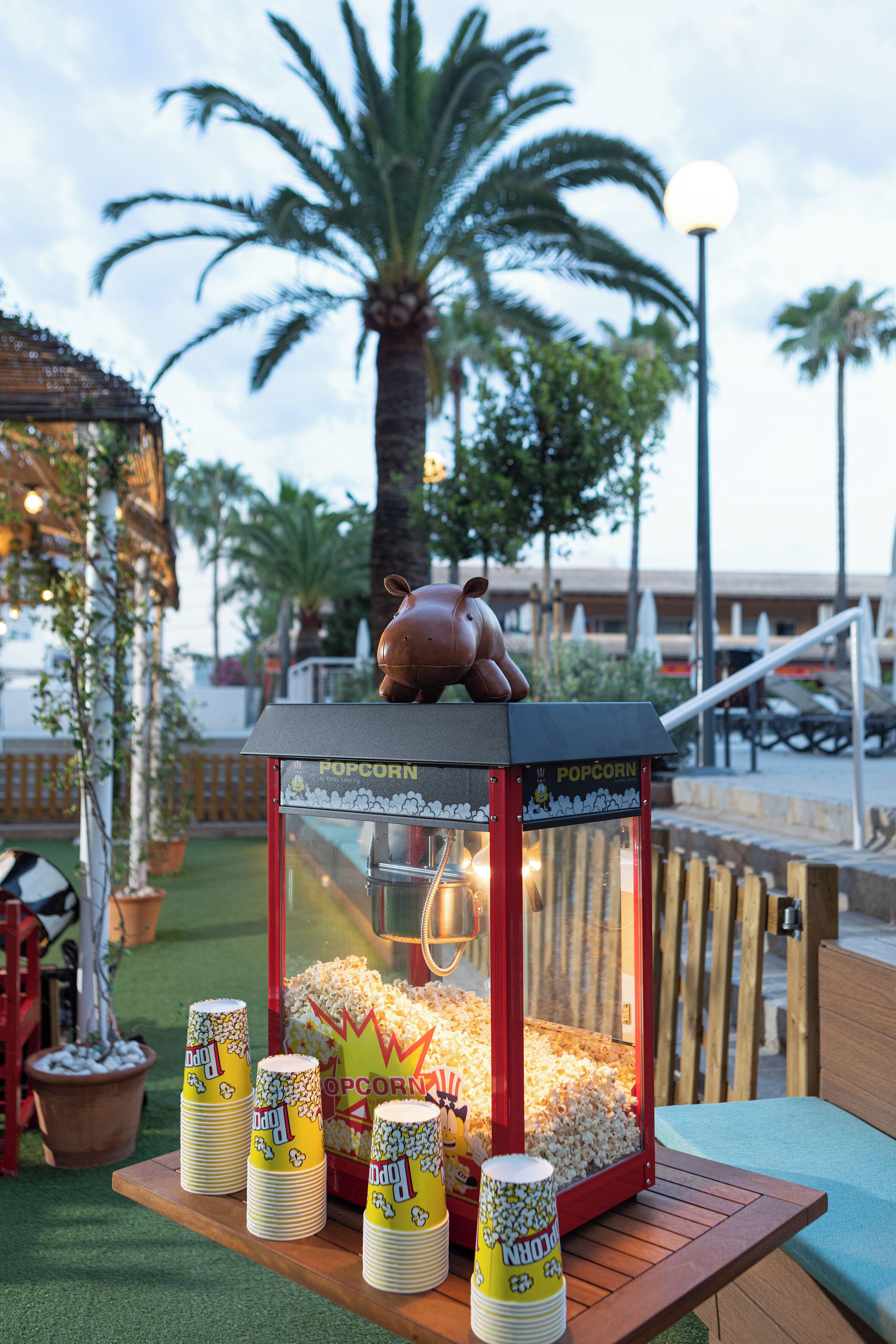Un stand de pop-corn surmonté d’une figurine de cochon, entouré de palmiers et doté d’une terrasse en bois.
