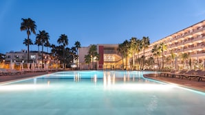 Un espace piscine d’hôtel avec des chaises longues et des palmiers, entouré d’immeubles dotés de balcons.