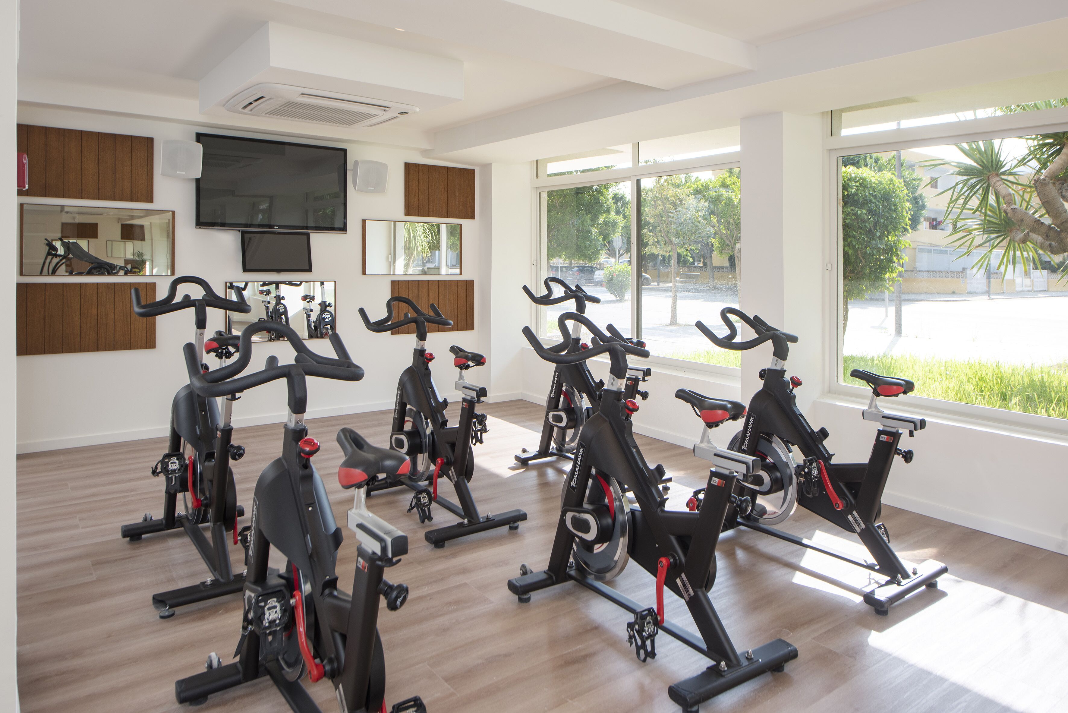 A gym with stationary bikes arranged in rows, large windows, and a flat-screen TV mounted on the wall.