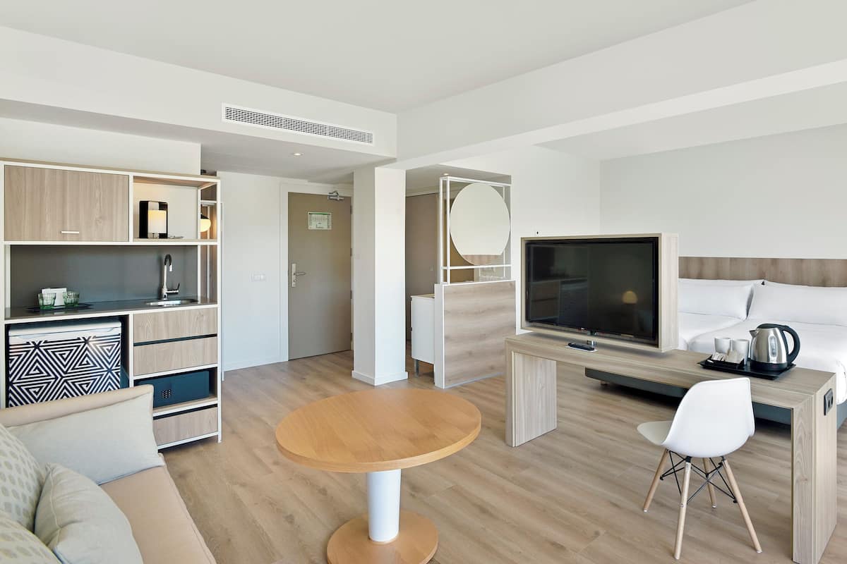 A modern hotel room with a kitchenette, a TV, a bed, a round wooden table, and a sofa.