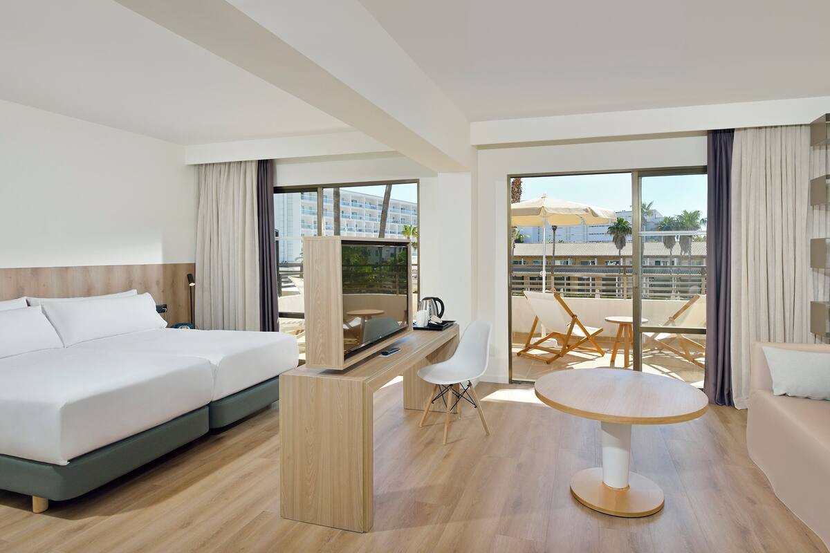 Une chambre d’hôtel moderne dotée d’un grand lit, d’un bureau avec une télévision, d’un balcon aménagé avec des sièges et offrant une vue sur le paysage urbain.