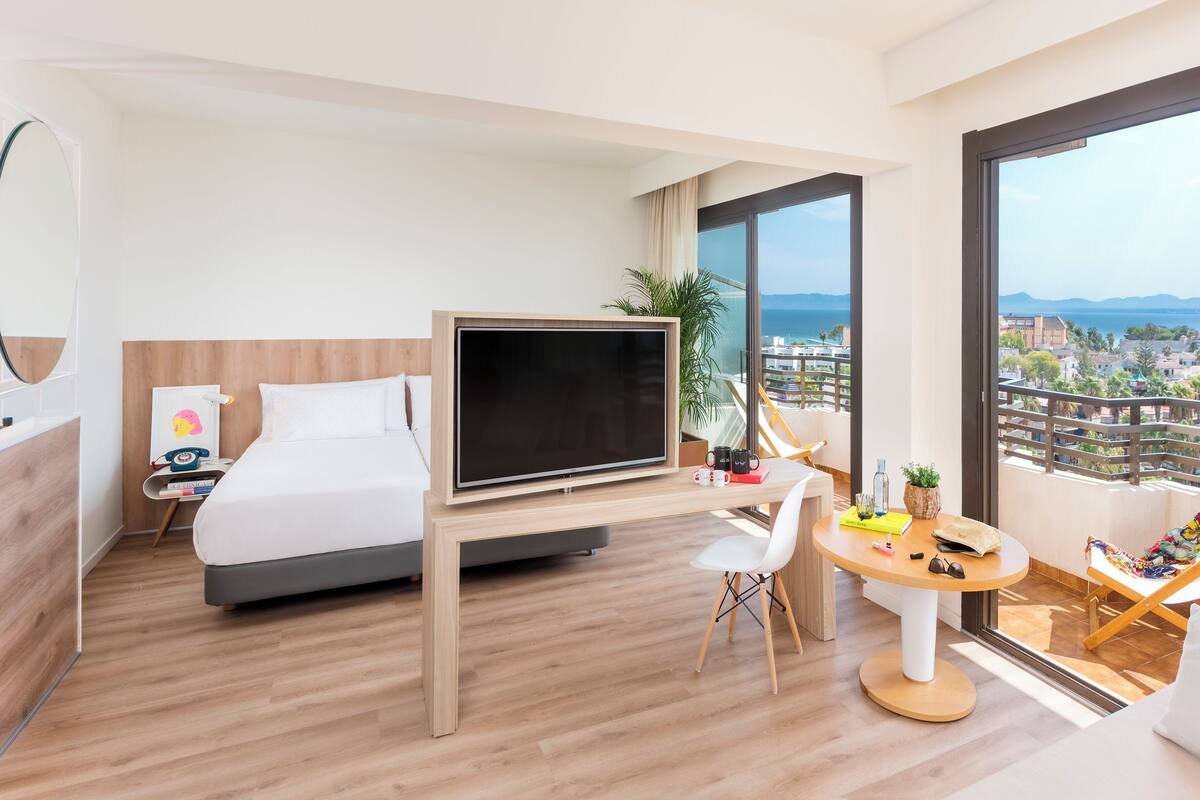 Une chambre d’hôtel moderne équipée d’un lit, d’une télévision, d’un bureau et d’un balcon offrant une vue sur la ville.