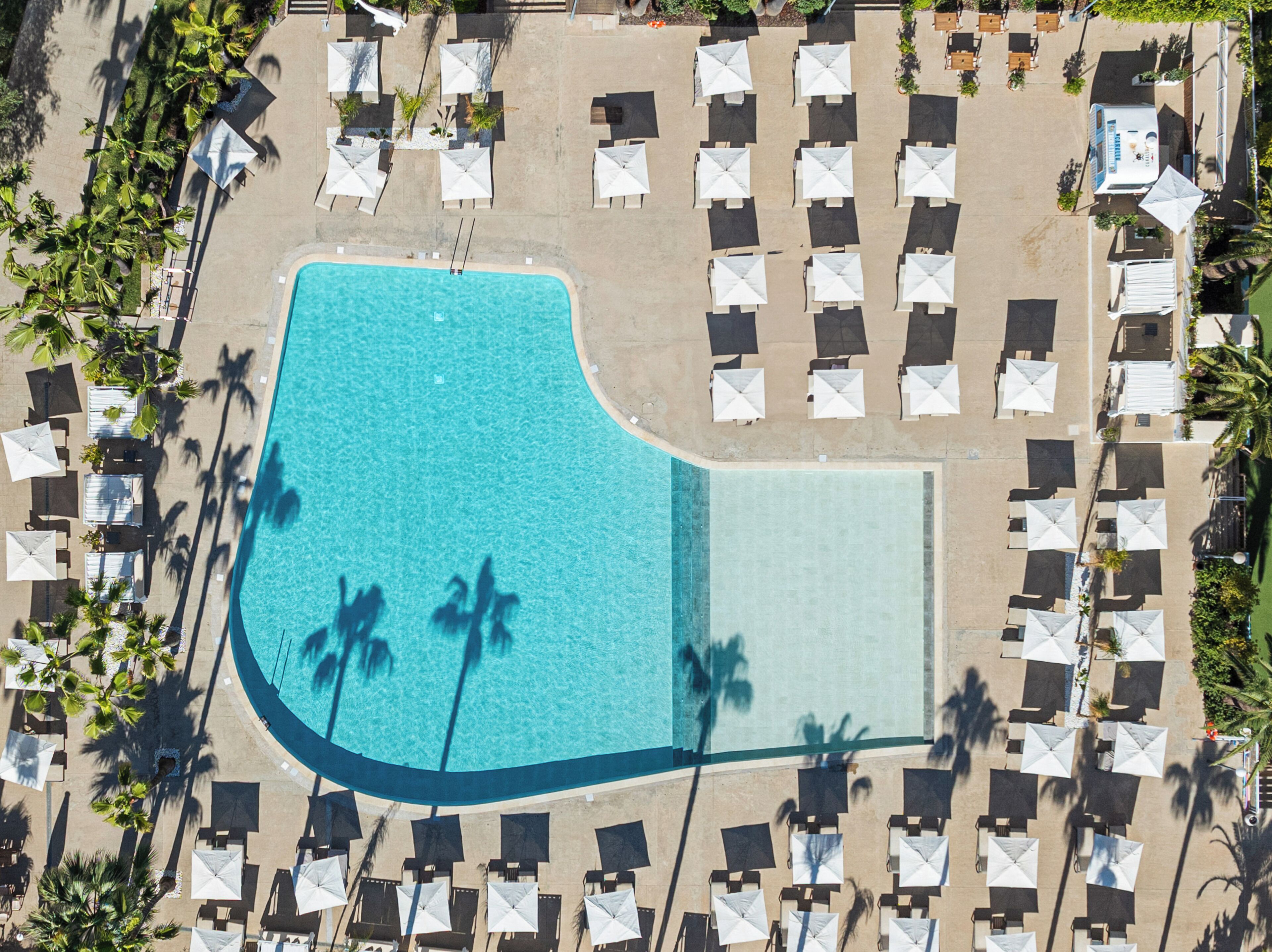 Vista aérea de un complejo turístico con una piscina grande, varias zonas de descanso con sombrillas y palmeras.