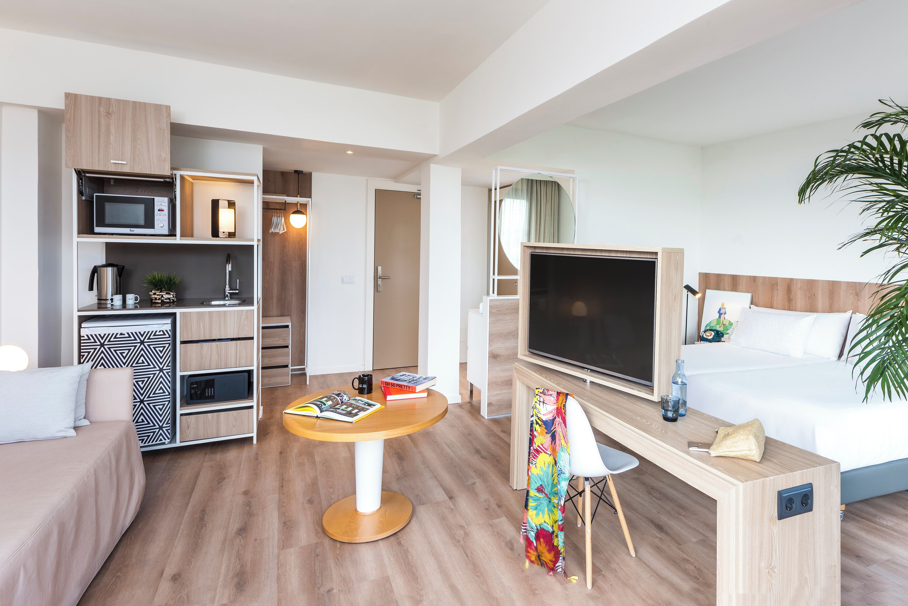 Ein modernes Hotelzimmer mit Bett, Fernseher, Schreibtisch mit Stuhl, einem kleinen Tisch und einer Küchenzeile.