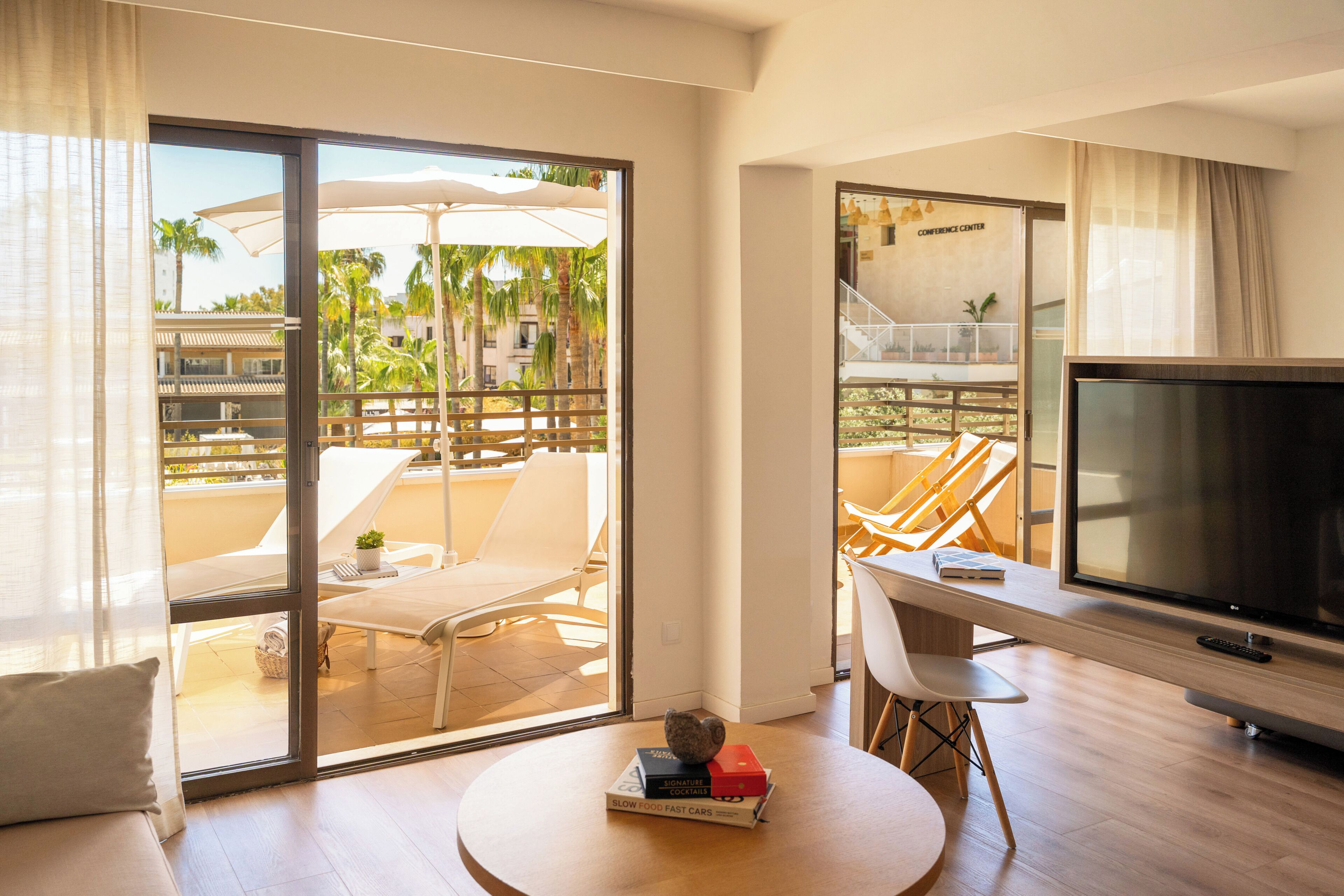 A modern hotel room with a balcony, a TV, a round table with books, and a view of a palm tree and building.