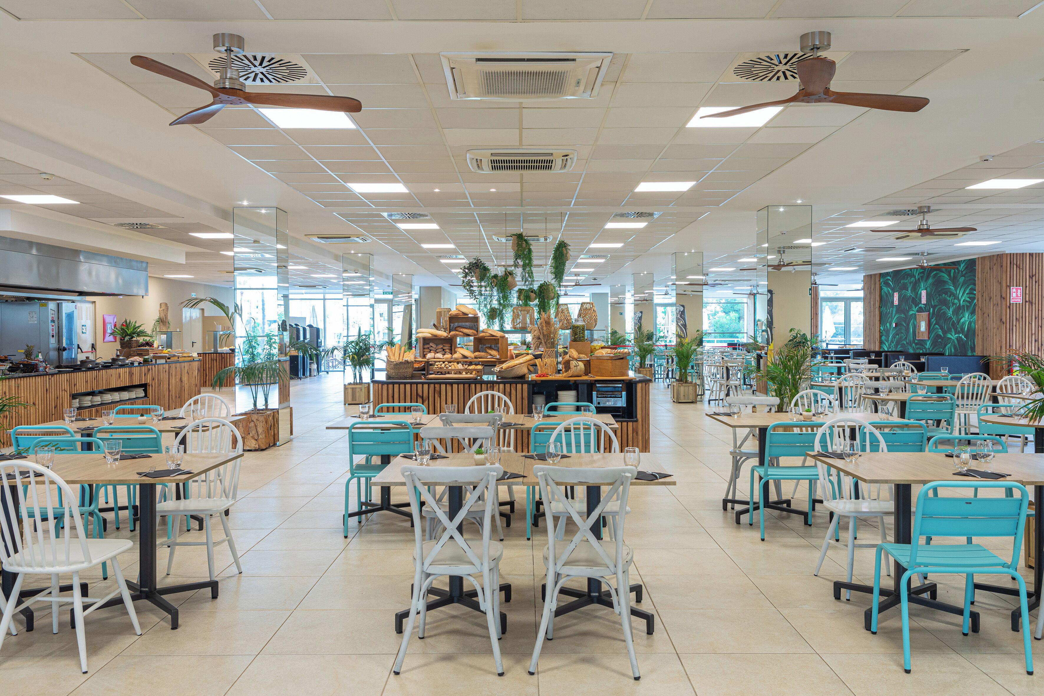 Een ruime, overdekte eetruimte met meerdere tafels en stoelen, plafondventilatoren en een centraal aanrecht met etalages.