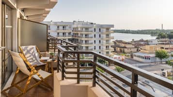 A balcony with a view of a river and buildings, wooden chairs, and a small table with a plant.