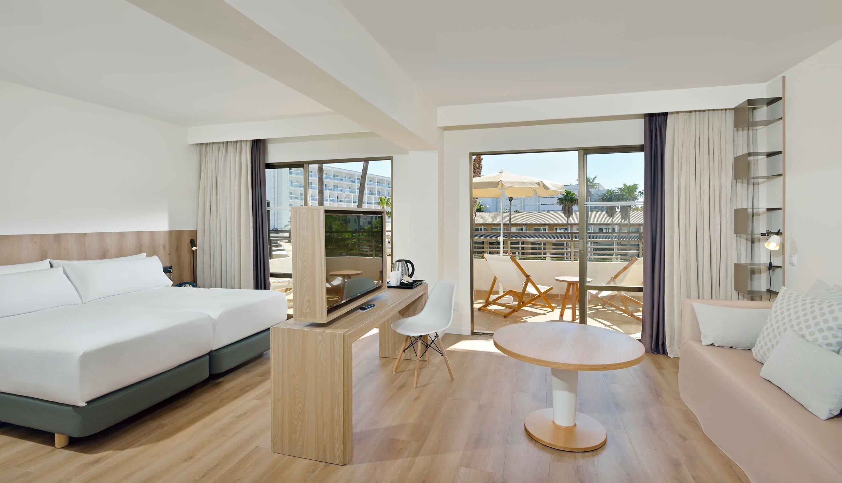 Ein Hotelzimmer mit Bett, Schreibtisch mit Fernseher, Balkon mit Tisch und Stühlen sowie einer Couch.