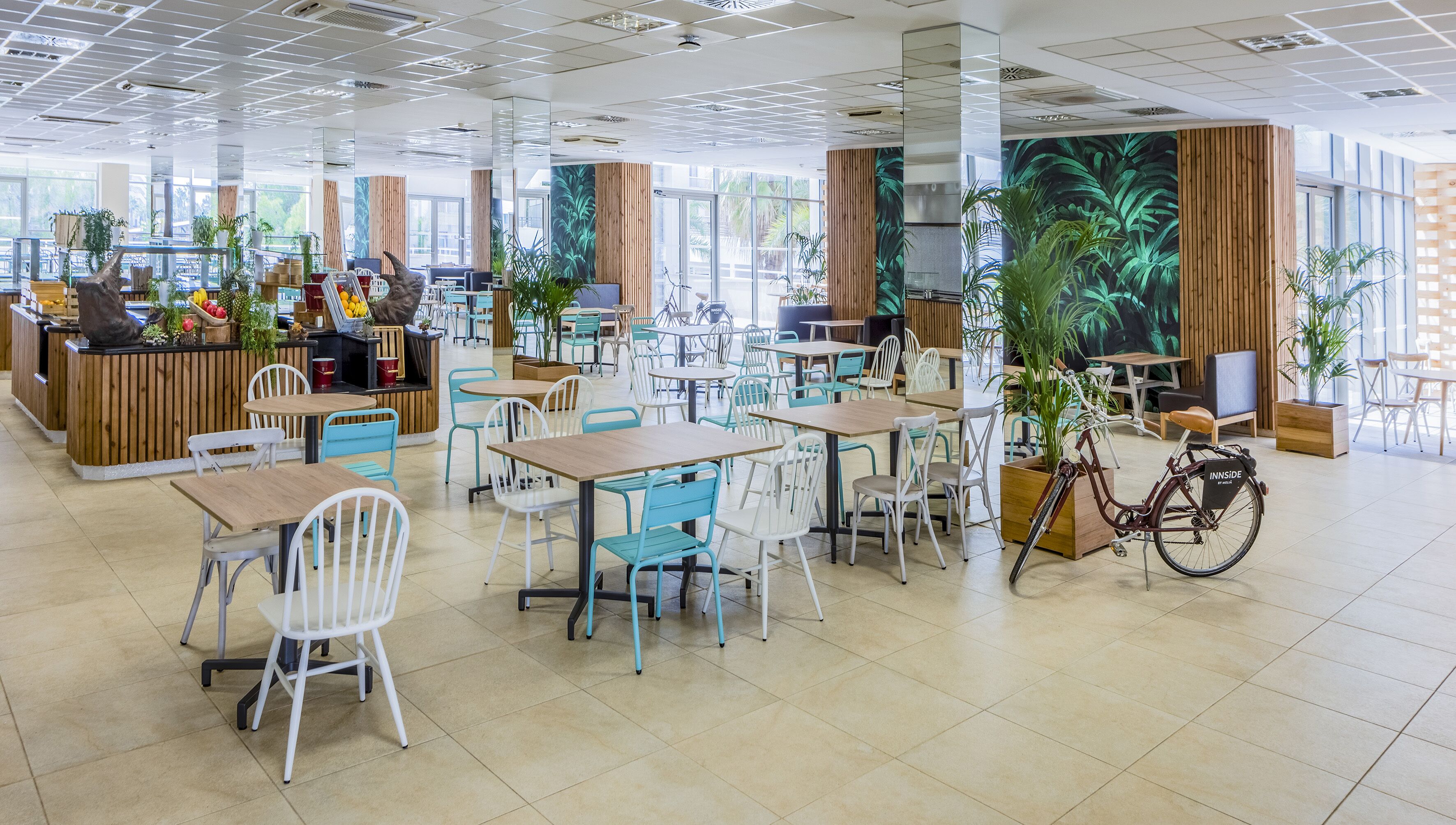 Un café amplio con varias zonas de estar, incluidas mesas y sillas, y una barra central con una exhibición de frutas y bebidas.