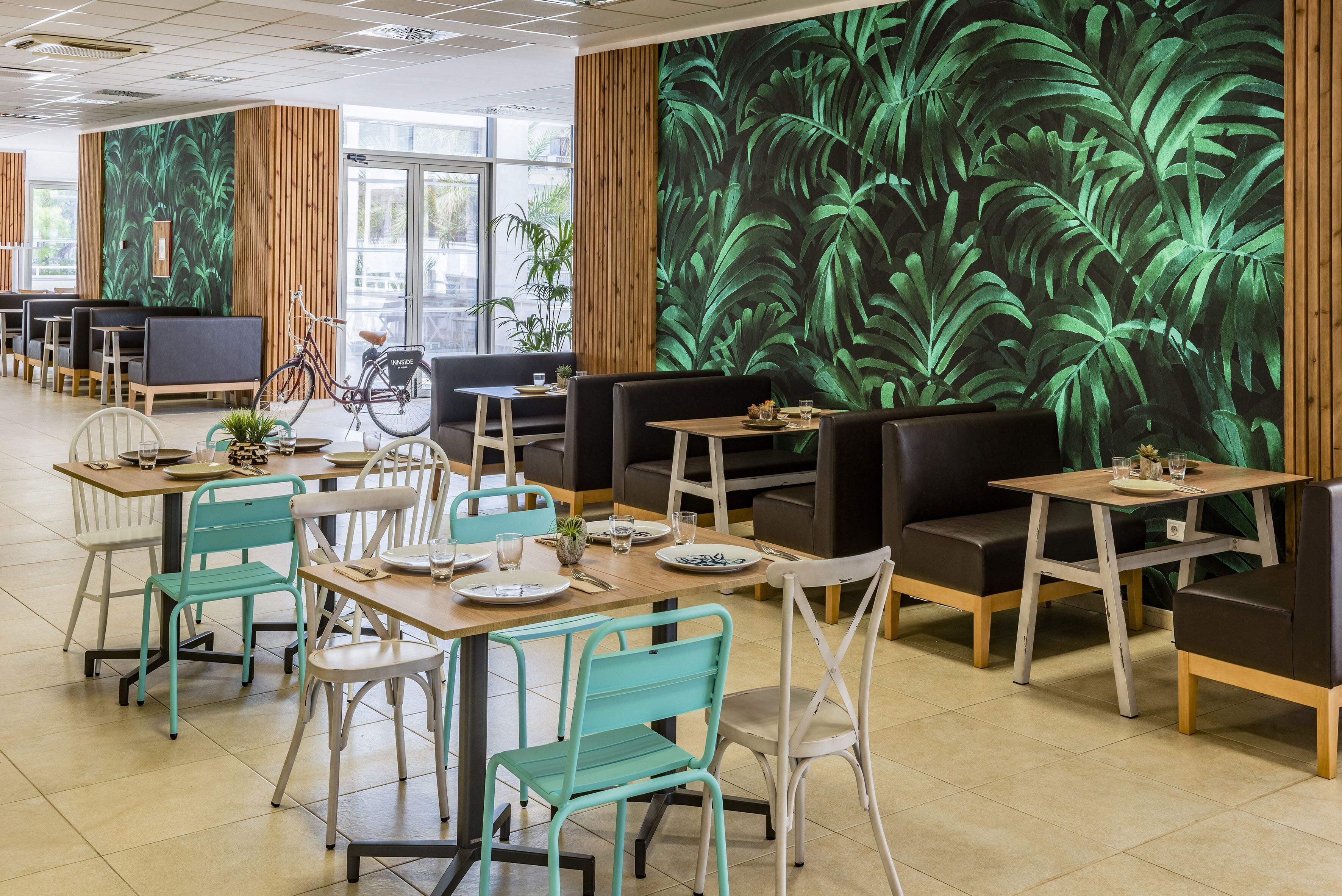 Un restaurant moderne avec des murs lambrissés en bois, des œuvres d’art murales représentant des feuillages verts et différents types de sièges, notamment des tables et des banquettes.