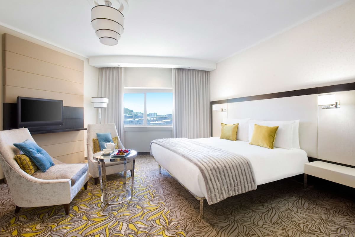 A hotel room with a bed, two armchairs, a small table, a TV, and a window with a city view.