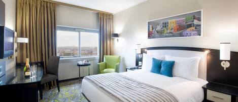 A hotel room with a large bed, a desk with a bottle of champagne, a chair, a small table, and a window with curtains.