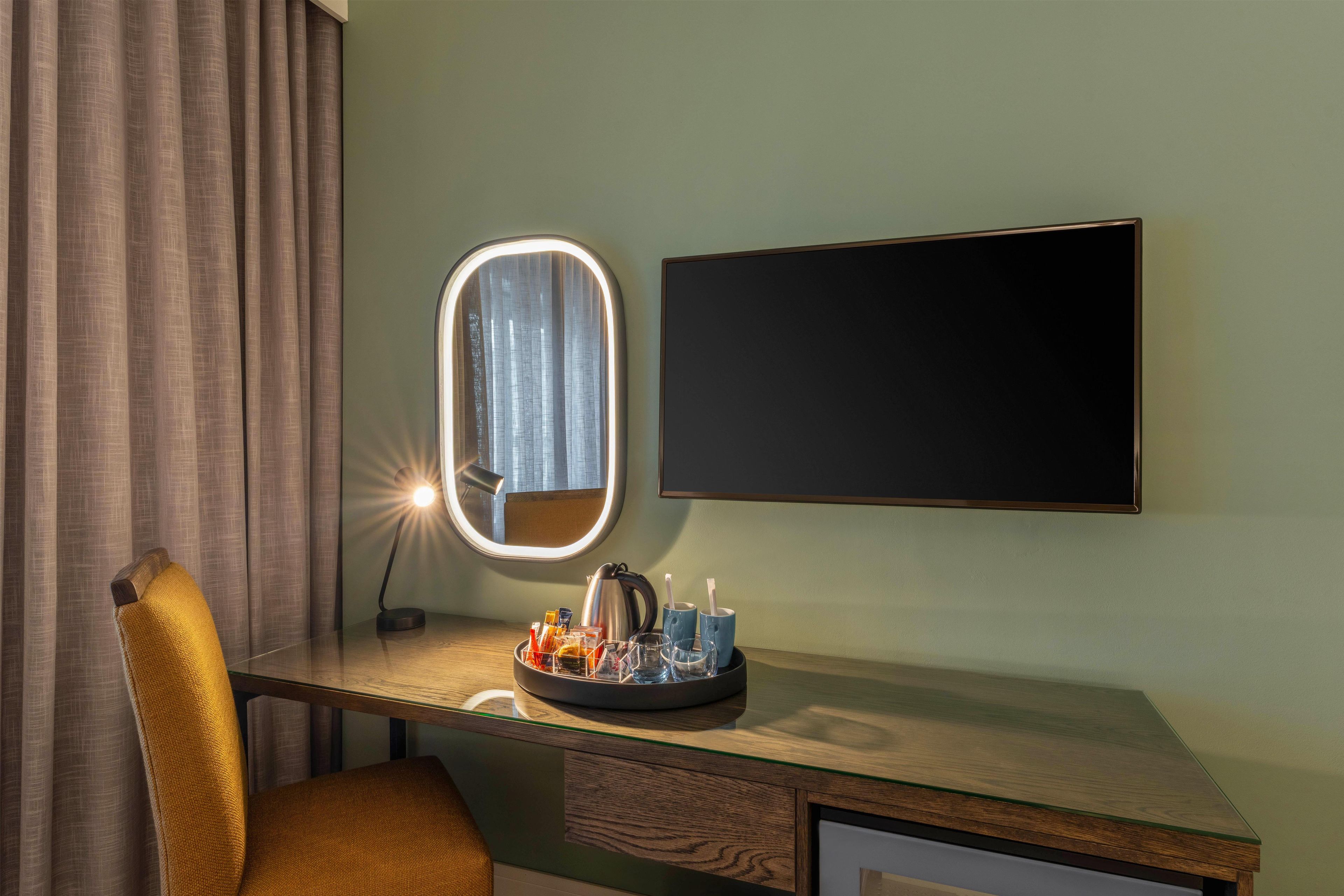 A hotel room with a vanity area, a mirror with lights, a flat-screen TV, and a desk with a coffee maker and glasses.