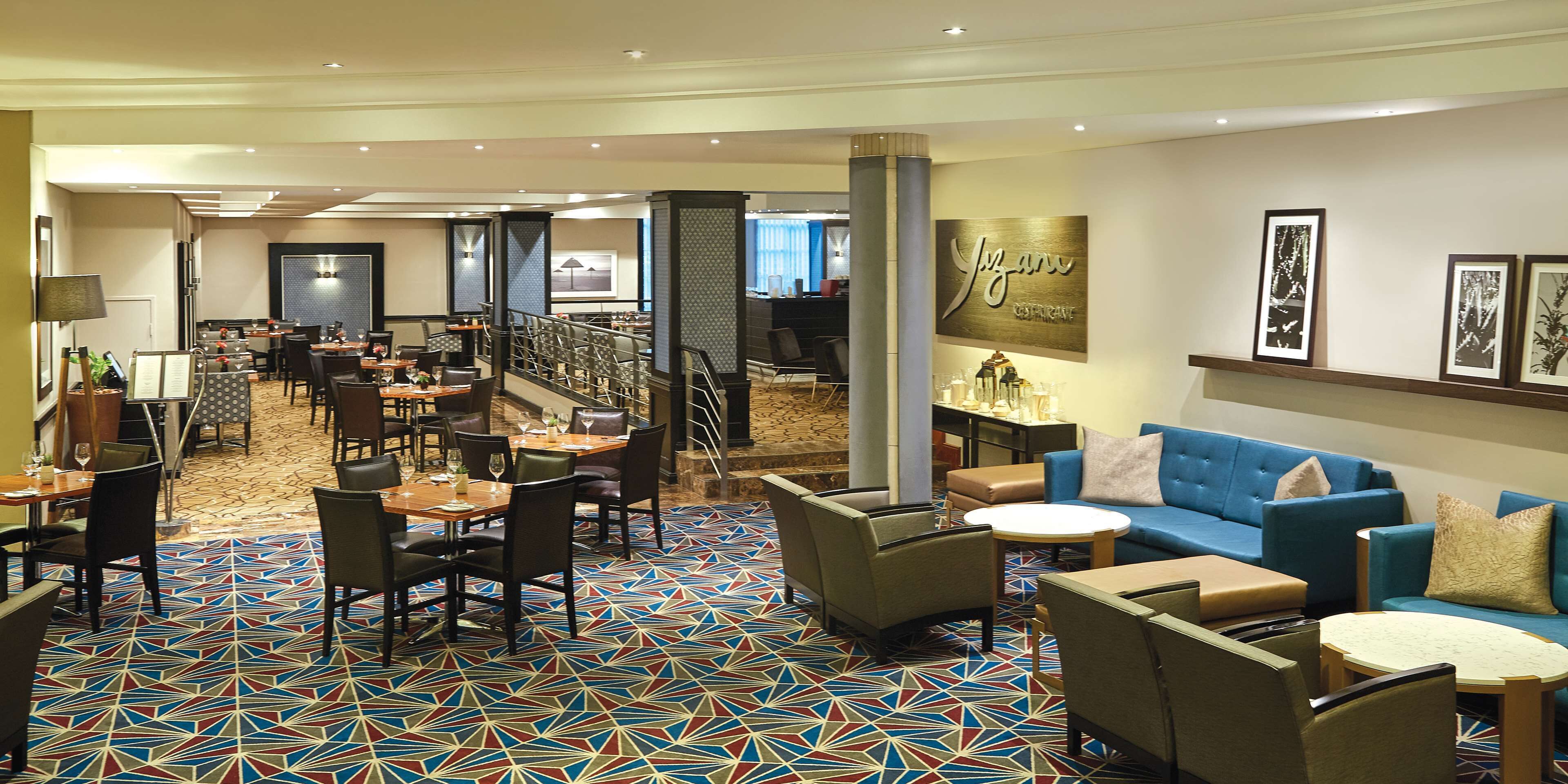 A hotel lobby with a patterned carpet, seating areas, a bar, and a wall with framed pictures.
